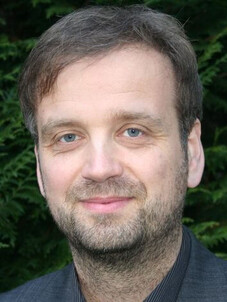 Oliver G. Schmidt vor grünem Laubhintergrund, dezent lächelnd.